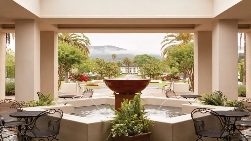 A peaceful outdoor courtyard features a round central fountain with water flowing into a basin. Surrounding the fountain are elegant, metal garden chairs and tables. The backdrop showcases palm trees, greenery, and misty mountains under a cloudy sky, ideal for guests at nearby Napa hotels.