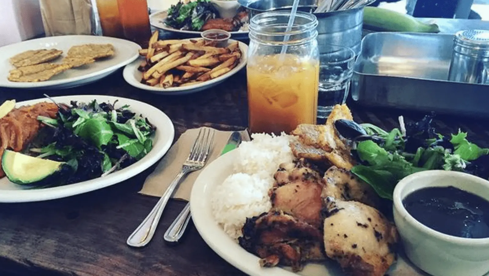 A table set with plates of food for the best lunch in North Bay: a main plate featuring grilled chicken, rice, salad, and a small bowl of sauce; another plate with fries; another plate with salad and proteins; a mason jar with iced tea, and additional condiments and metal trays in the background.