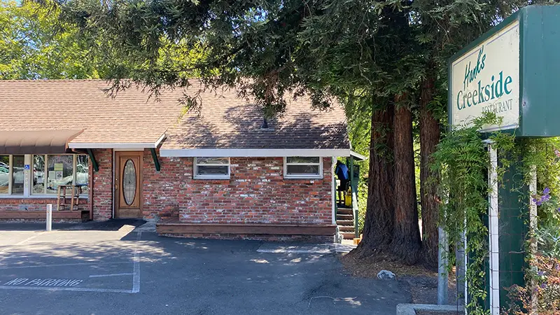 Brick building with a sign reading "Fork's Creekside" surrounded by tall trees. The building has a brown shingled roof, an oval glass entry door, and several windows. "No Parking" sign painted on the ground.