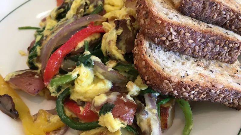 A plate of scrambled eggs mixed with red and green bell peppers, red onions, and spinach served alongside two slices of toasted multigrain bread. The food is arranged on a white plate with a green rim.