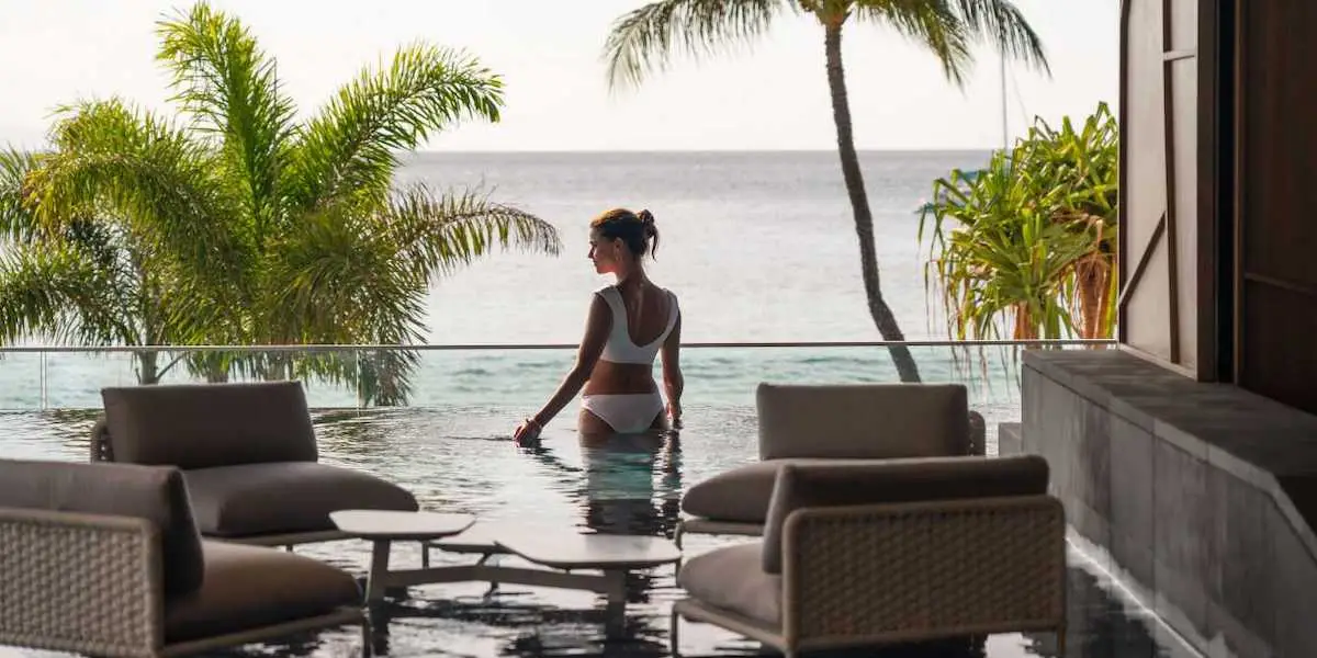 A woman in a white swimsuit stands in an infinity pool facing the ocean, with palm trees and patio furniture in the foreground. The scene is serene and tropical, suggesting a luxurious and relaxing setting at one of Hawaii's finest hotels with spas.