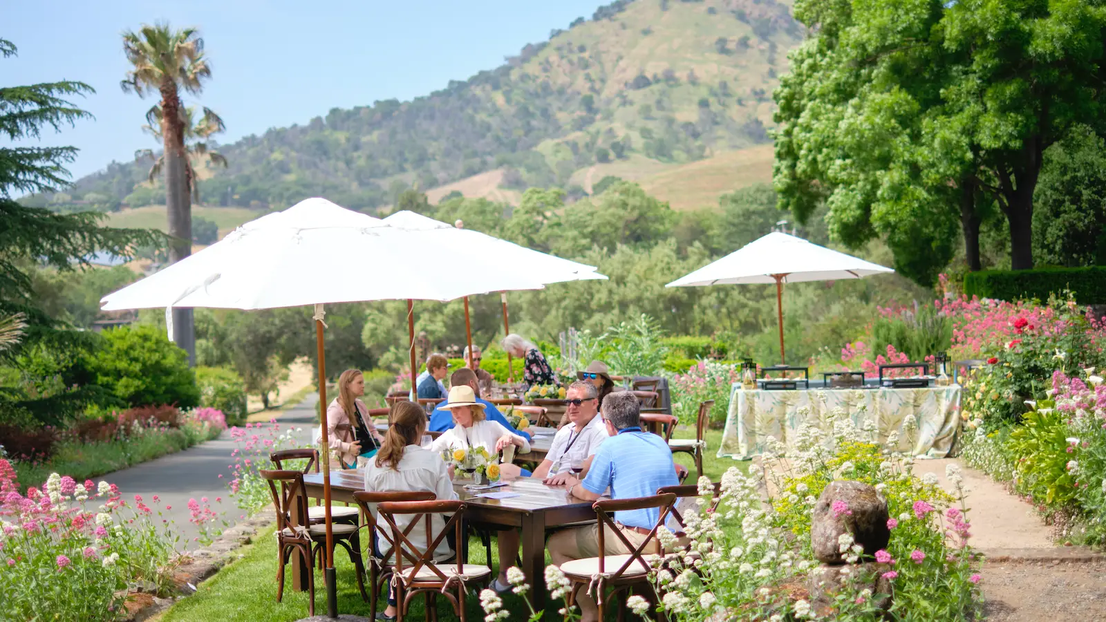 A group of people sitting at a wooden outdoor table under white umbrellas in a garden setting. They are surrounded by colorful flowers and greenery, with a scenic hillside in the background. Tables are decorated, and the scene is bright and sunny—one of the best things to do in Napa in April.