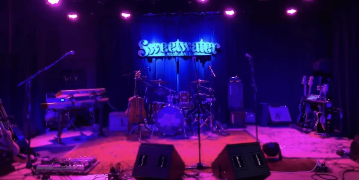 A dimly-lit music stage with purple lighting awaits in North Bay this September. The setup includes a drum kit, keyboards, amplifiers, and various microphones. Above the stage, a lit sign spells "Sweetwater." The scene is ready for a live performance but currently empty of musicians—one of the best things to do this month.