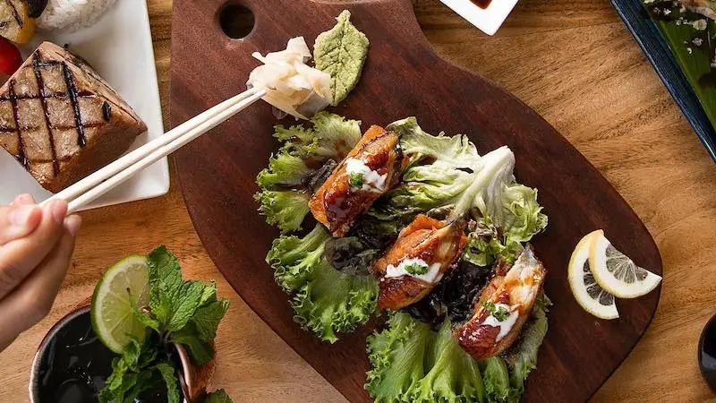 A person is using chopsticks to pick up ginger from a small pile on a wooden board. The board also has three pieces of grilled meat wrapped in lettuce leaves, garnished with a green sauce. Nearby, there are lemon slices, mint leaves, and other dishes—all set against the picturesque backdrop of a Sunset Dinner in Kauai.