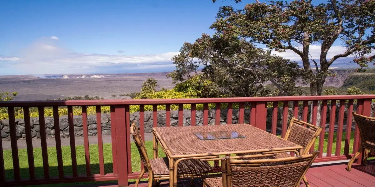 A wooden deck with a table and chairs overlooks a scenic landscape featuring a vast expanse of land, trees, and mountains in the background. Enjoying the best lunch in Hilo here would be perfect under the clear blue sky, enhancing the serene atmosphere of the view.