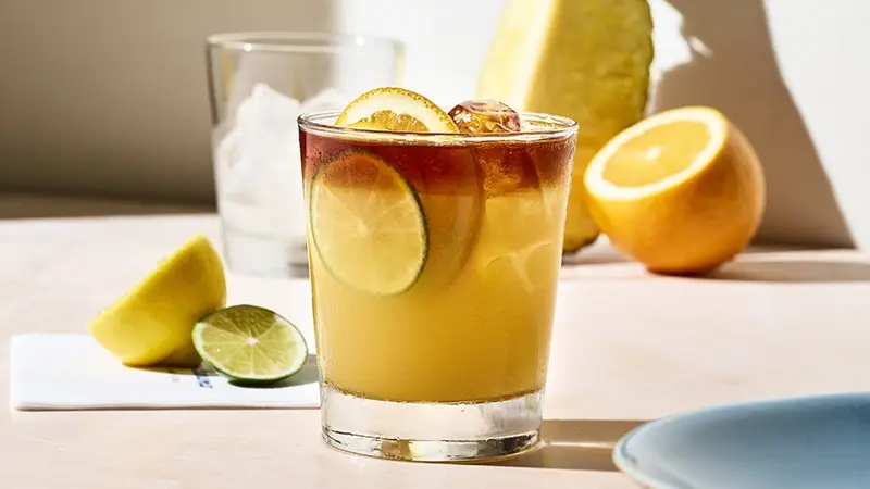 A glass filled with an iced drink garnished with lime and lemon slices sits on a sunny table. Behind it, there are lemon and lime halves, a whole orange, and a clear glass containing ice cubes. The setting, reminiscent of the best happy hour on the Big Island, is bright with natural light.