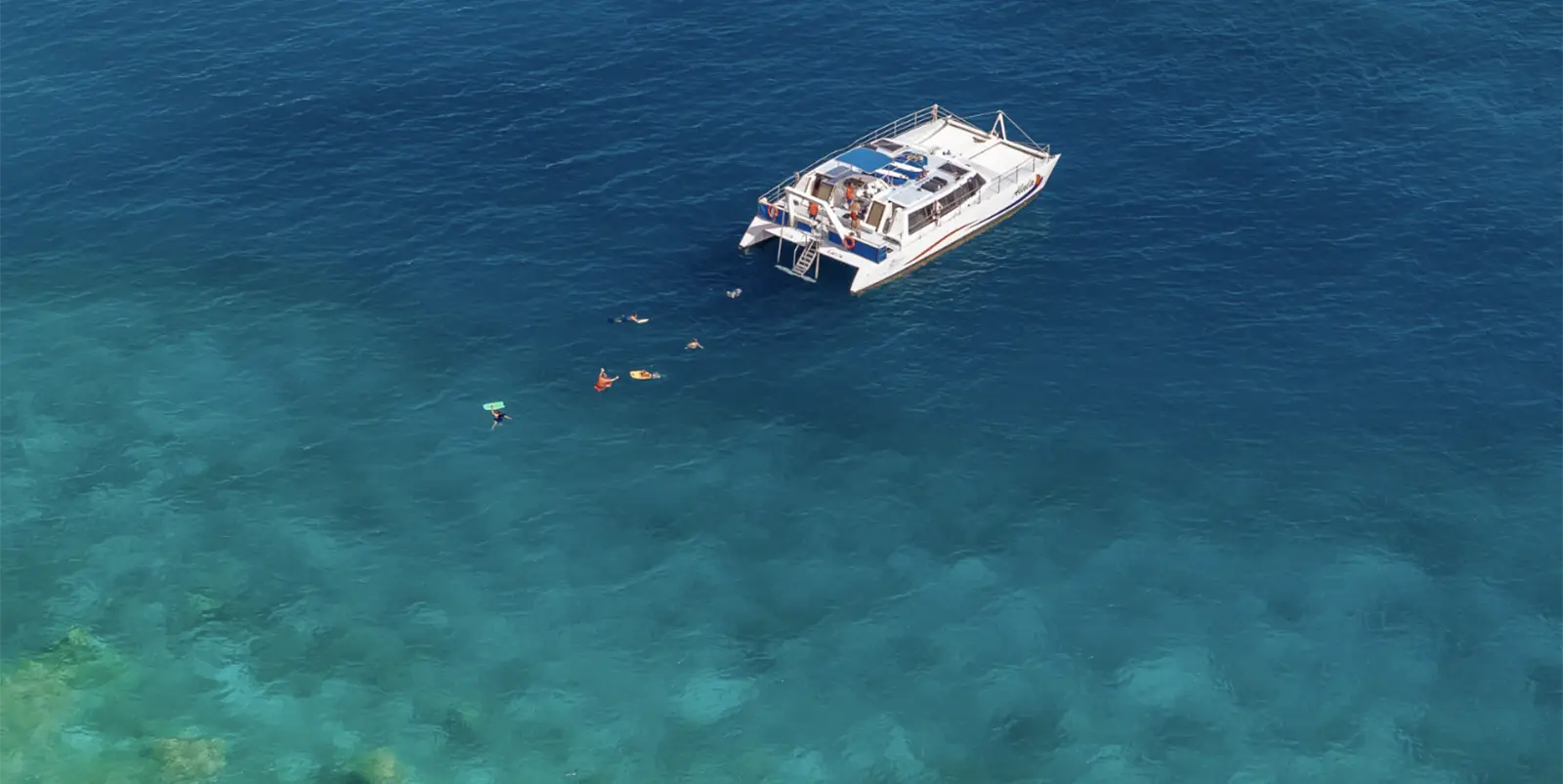 Aerial view of a large white catamaran floating on clear blue water. Four people are swimming near the boat, enjoying the ocean—one of the top water activities on the Big Island. The water transitions from a lighter turquoise near the bottom of the image to a deeper blue further away.