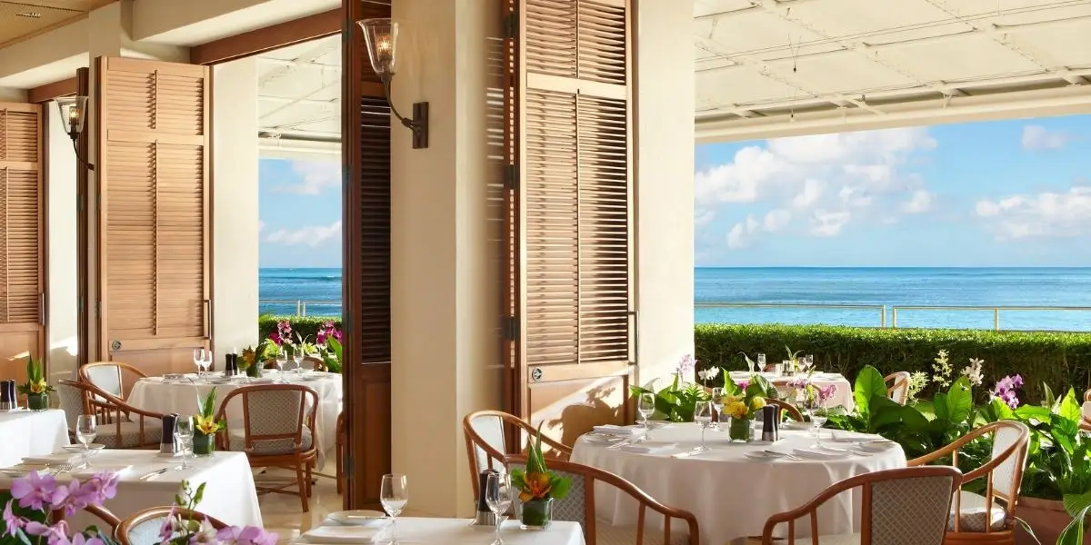 An elegant seaside restaurant at Halekulani Hotel Waikiki features tables set with white linens and floral centerpieces. Wooden shutters are opened wide, revealing a view of the ocean and a sunny, blue sky. The atmosphere is serene and inviting, perfect for a scenic dining experience.