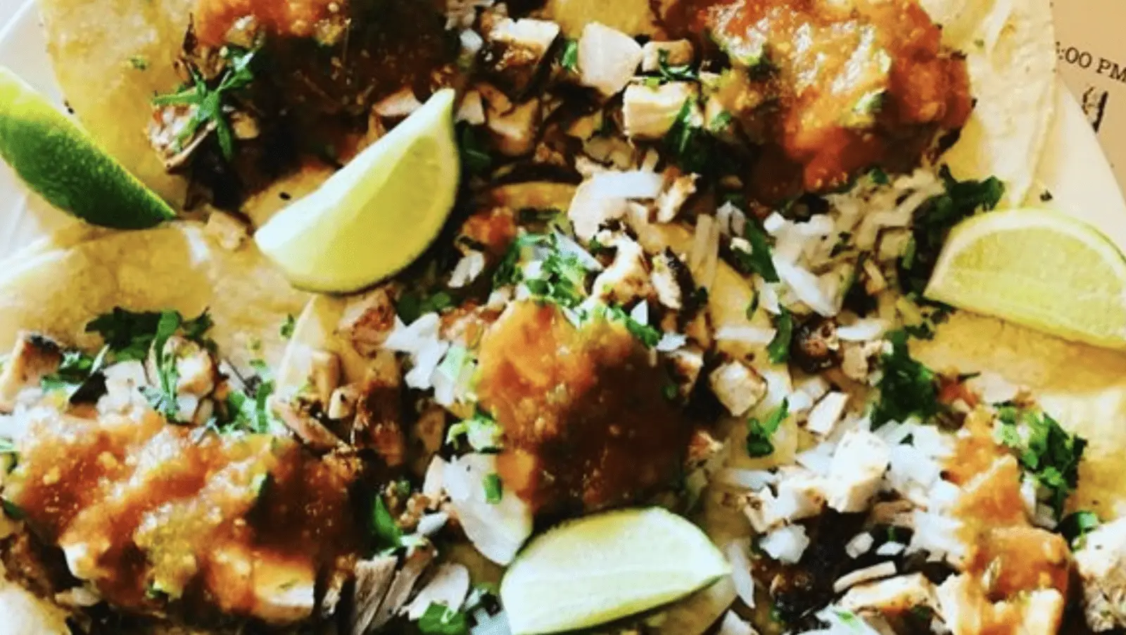 A close-up image of chicken tacos garnished with diced onions, cilantro, and salsa, topped with lime wedges, on a plate. The tacos have a vibrant mix of colors from the greens of the cilantro and limes, the whites of the onions, and the reddish-brown salsa.