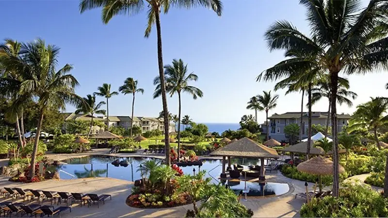 A picturesque tropical resort, hailed as one of the Best Hotels on Kauai, features a central swimming pool surrounded by lounge chairs and umbrellas, lush gardens, and tall palm trees. In the background, there are resort buildings and a view of the ocean under a clear blue sky.