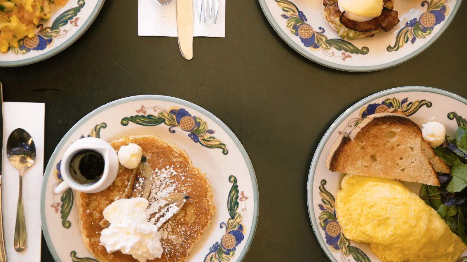 Four plates of food on a dark green table. The plates contain pancakes with syrup and whipped cream, an omelet with toast, and various other breakfast items. Utensils are placed next to some of the plates. The plates have a floral pattern.