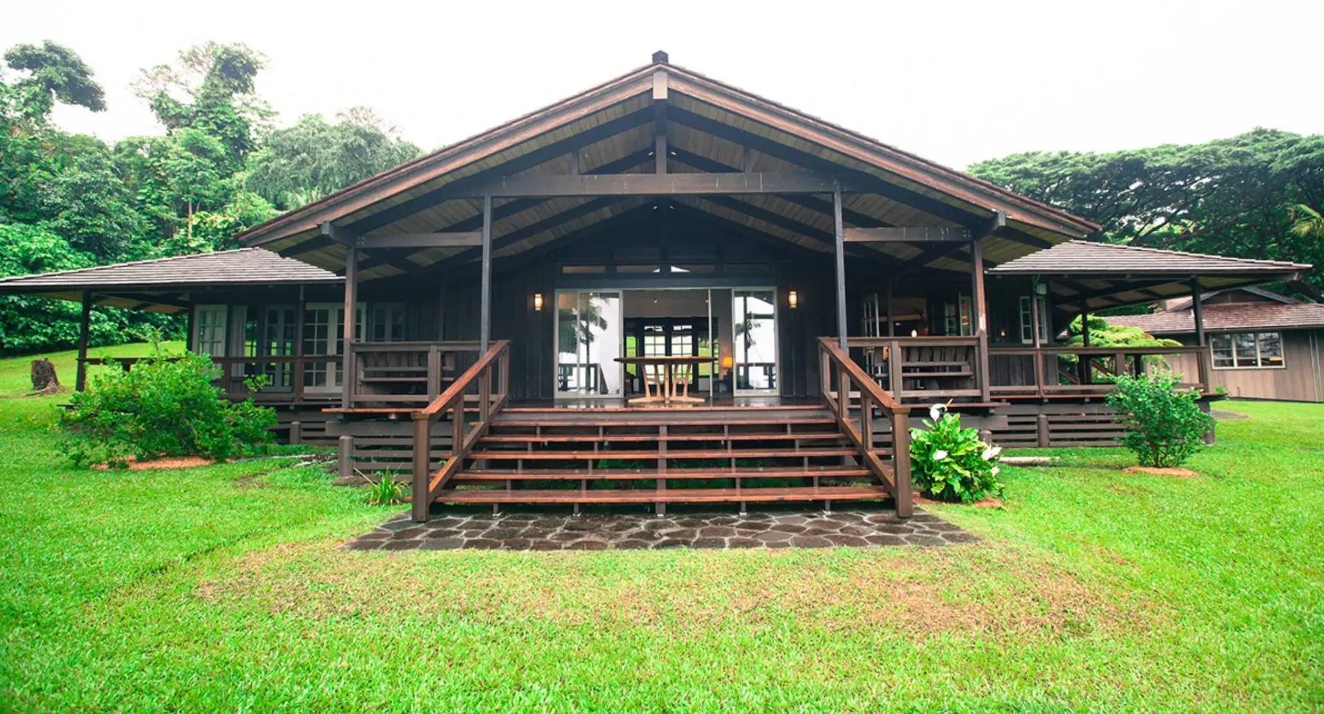 A cabin-style house with a large front porch, wooden steps leading up to the entrance, and a lush green lawn. The structure features a high triangular roof and large windows, much like the serene retreat options you'd find at the best group hotels in Maui, nestled in a tropical forested area.