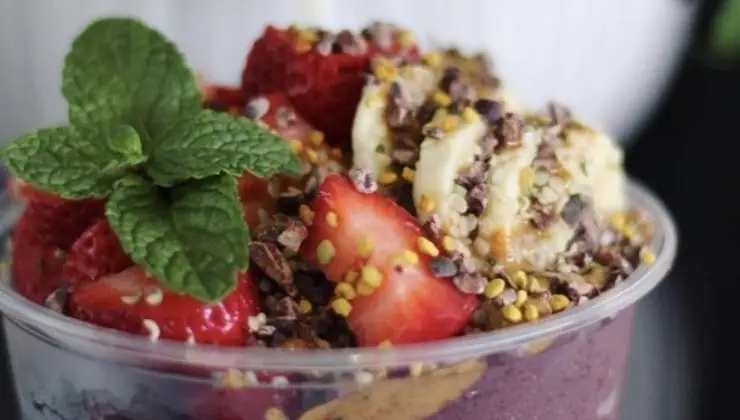 A close-up of a colorful acai bowl topped with fresh strawberries, banana slices, cacao nibs, bee pollen, and a sprig of mint. The smoothie base appears to be purple, likely made from berries or acai—the presentation is reminiscent of the best smoothies on Hawaii Island.
