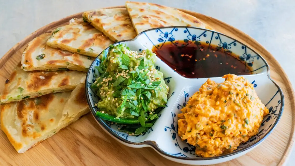 A wooden plate holds crispy, golden scallion pancakes cut into triangular slices, perfect for the best dinner in Wine Country. Beside the pancakes, a ceramic dish with three compartments contains green avocado salad garnished with sesame seeds, a brown dipping sauce, and a creamy orange dip.