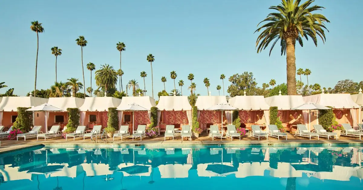 A luxurious outdoor swimming pool, one of the best hotel pools in California, is surrounded by lounge chairs and umbrellas in front of a row of cabanas. Tall palm trees and clear blue skies complete the serene, tropical atmosphere.