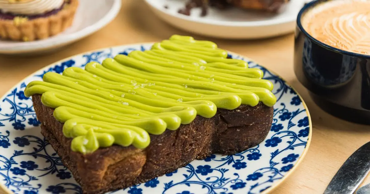 A slice of toast covered with green frosting, shaped like a Christmas tree, sits on a blue and white plate. The plate is on a wooden table next to a cup of coffee and a partially visible dessert in the background.