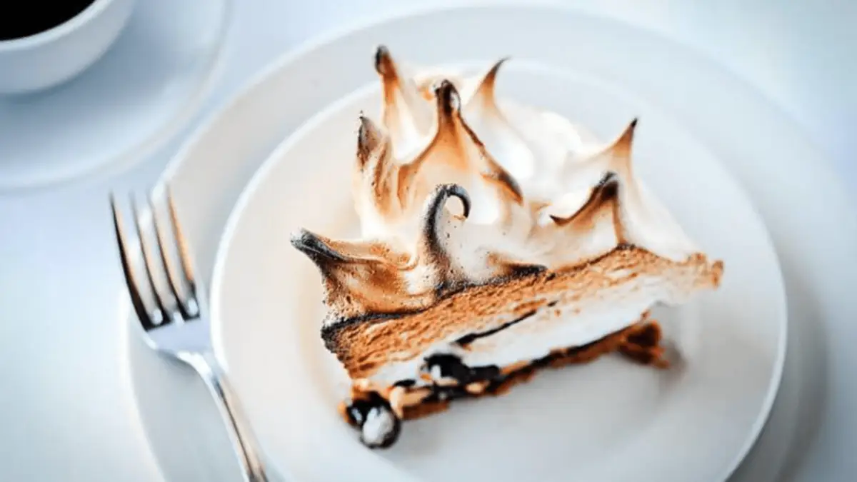 A slice of baked Alaska dessert from one of the Best Desserts in the Bay Area rests on a white plate. The dessert has browned peaks of meringue on top, covering a layer of cake and ice cream. A fork is placed on the left side, ready for use. The background is softly blurred with a hint of a coffee cup.