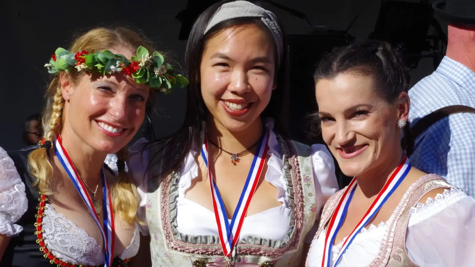 Three smiling women, dressed in traditional attire and adorned with medals, pose together outdoors. The woman on the left sports a floral crown, the one in the middle wears a headband, and the woman on the right has her hair tied back. It's one of the best things to do in the Bay Area October festivities.