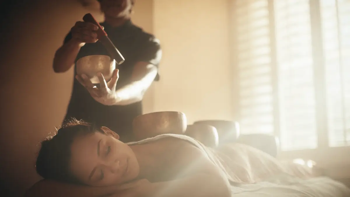 A person lies face down on a massage table with eyes closed, appearing relaxed. Another individual holds a singing bowl above the person’s back in one of the Best Spa and Wellness Hotels in the Bay Area. Soft light filters through a window, creating a serene and peaceful atmosphere.