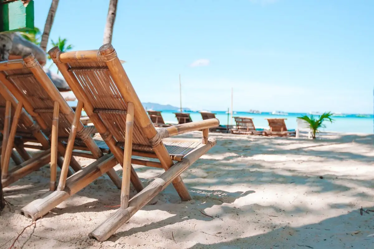 Two wooden lounge chairs are arranged on the sandy Poipu beach with additional chairs in the background. The turquoise sea extends to the horizon under a clear blue sky, and a few palm trees provide partial shade over the chairs.