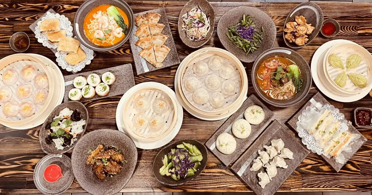 A wooden table filled with various Asian dishes, including steamed dumplings, soup, salads, fried appetizers, stuffed buns, and assorted vegetables. Plates and bowls of colorful food are artistically arranged, creating an appetizing display—the best lunch San Francisco has to offer.