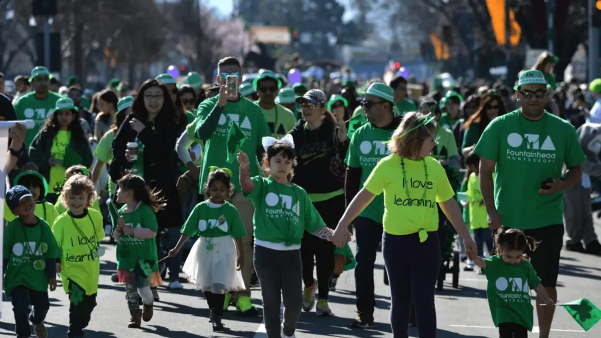 A joyful crowd of people, including adults and children, walk together in an outdoor parade. Most are wearing bright green shirts with a logo on them, and some have festive accessories like headbands and hats. They look happy and engaged in one of the Best Annual Events in the East Bay.