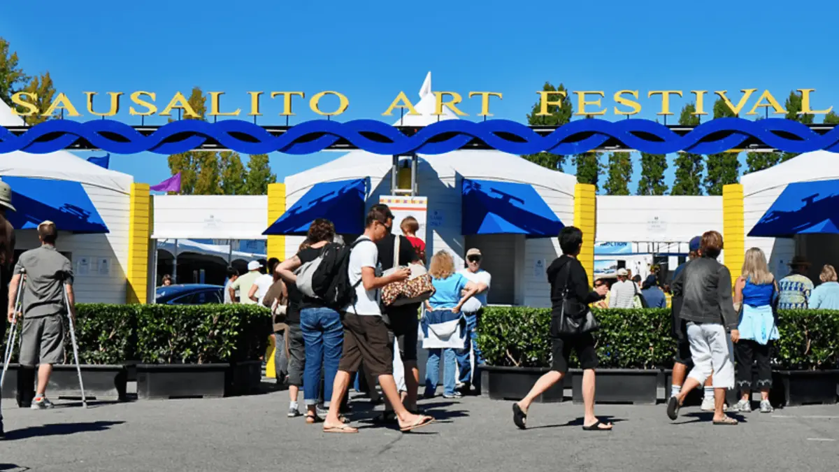 People walk towards the entrance of the Sausalito Art Festival on a sunny day. A large sign reading "Sausalito Art Festival" is displayed above, with decorative blue swirls below the text. As one of the best annual events in the North Bay, festival tents and booths are visible in the background.