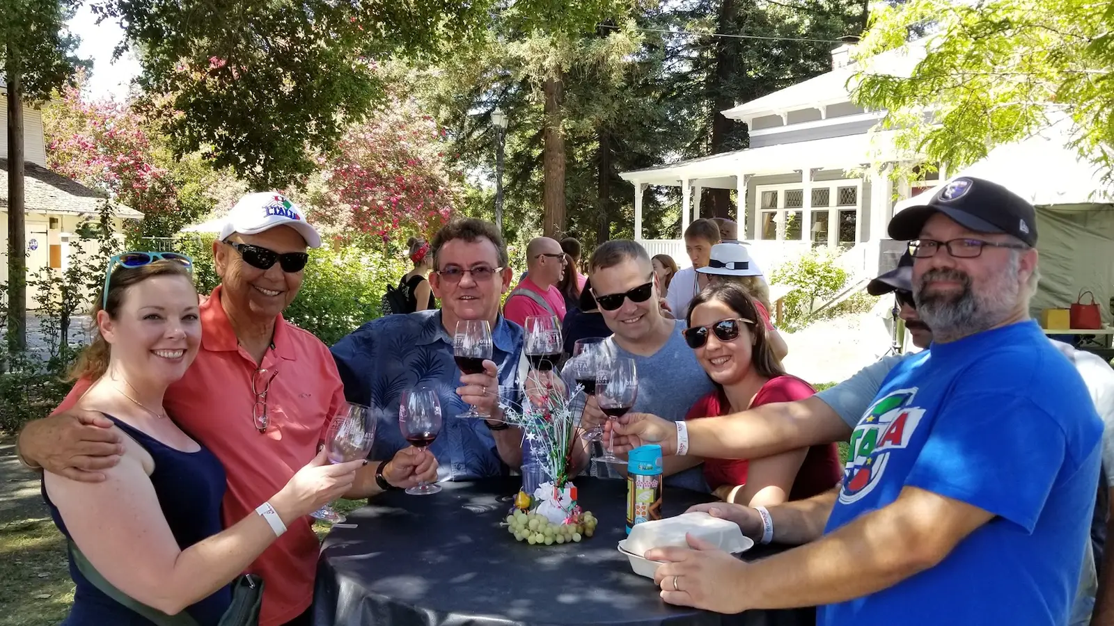 A group of seven people stands around an outdoor table, holding glasses of wine and smiling at the camera. They are in a garden setting with trees and a white house in the background. Casually dressed, they enjoy a sunny day at what appears to be one of the best things to do in the South Bay this August.