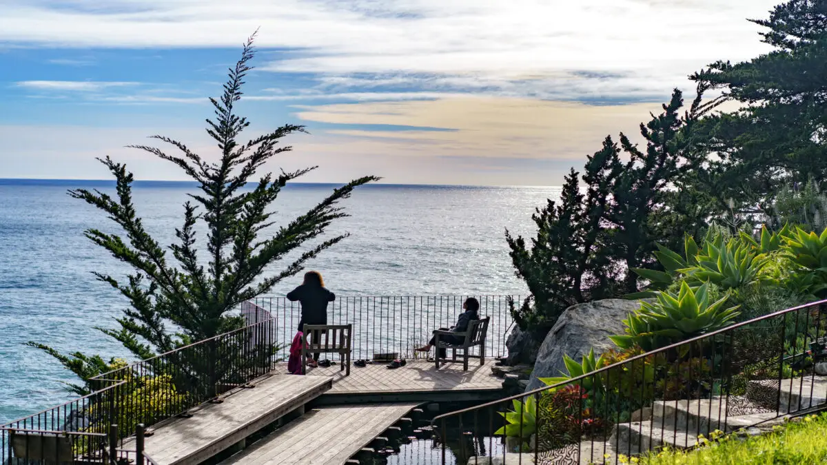 Two people enjoying a scenic ocean view from a wooden deck at Esalen, surrounded by lush greenery. One is standing and taking a photo, while the other is seated on a chair. Both are overlooking the calm, expansive sea with a cloudy sky in the background.