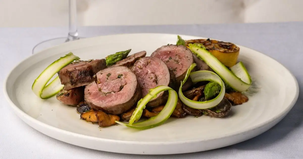 A gourmet dish featuring slices of rolled meat, garnished with thin asparagus spears, served on a bed of sautéed mushrooms. A charred lemon half is also placed on the plate for added flavor. The meal, one of the best dinners in San Francisco, is presented on a white plate with a drink in the background.