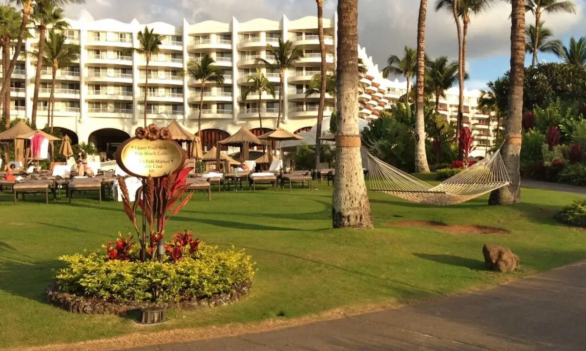 A luxurious resort scene with the grand, multi-story Fairmont Kea Lani in the background, surrounded by palm trees. In the foreground, a grassy area with lounge chairs, a hammock, and a tropical flower arrangement featuring a sign offers guests the perfect spot to relax and unwind.
