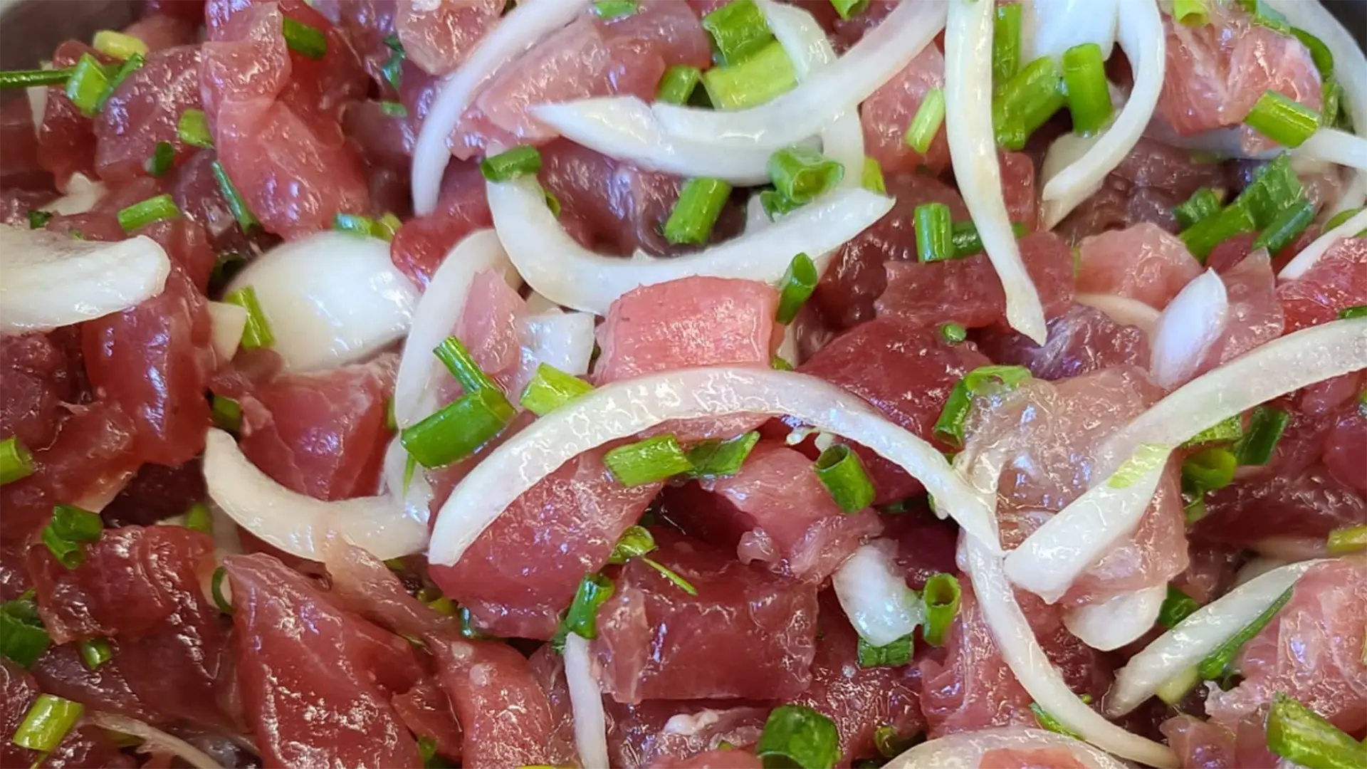 Close-up of a dish featuring raw pieces of tuna marinated with thin slices of onion and garnished with chopped green onions. The vibrant colors and fresh ingredients suggest a dish like poke or ceviche, reminiscent of the best poke in central Kauai.