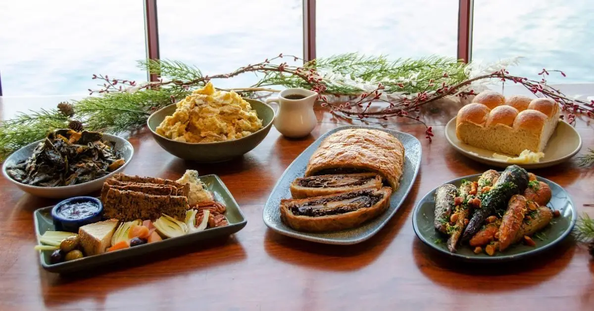 A table is set with a variety of dishes, including a loaf of bread, a plate of roasted carrots, slices of stuffed bread, a bowl of leafy greens, mashed potatoes, rolls, and a pitcher of sauce. The background features greenery and a window—truly the best lunch San Francisco has to offer.