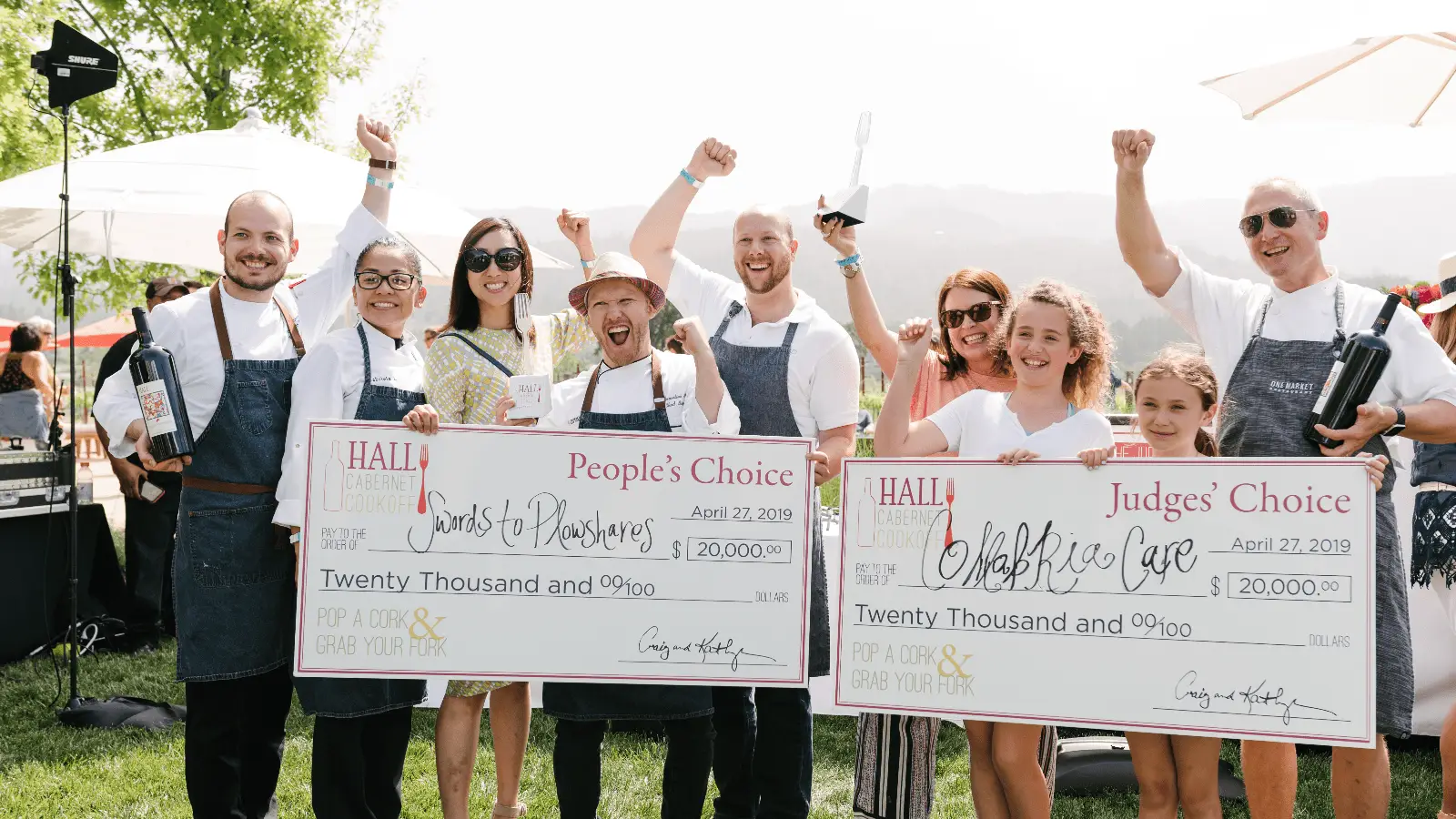 A group of smiling people stands outside, holding two oversized checks. One check reads "Sands of Thyme, $20,000" and the other reads "Garlicky Lads, $20,000." The group appears to be celebrating triumphantly—one of the best things to do in Napa this April.