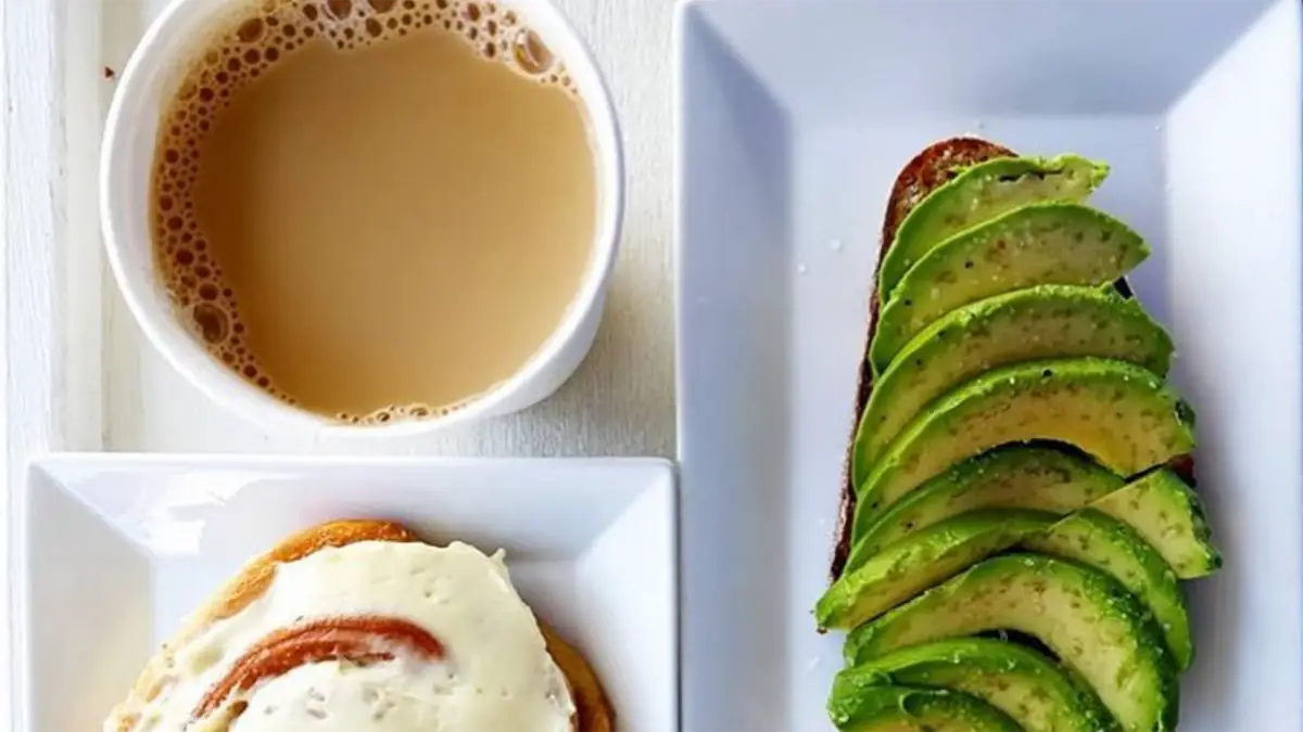 An overhead view of the best breakfast on Kauai's North Shore featuring a cup of coffee with cream, a plate with a cinnamon roll topped with icing, and a plate with sliced avocado on toast. The dishes are arranged on a white table.