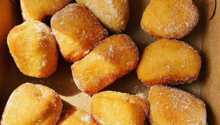 A close-up view of a batch of golden-brown, sugar-coated doughnuts arranged in a cardboard box. The best malasadas doughnut on Kauai, these treats have an irregular shape and a light dusting of sugar on their surfaces, suggesting a fresh and warm appearance.