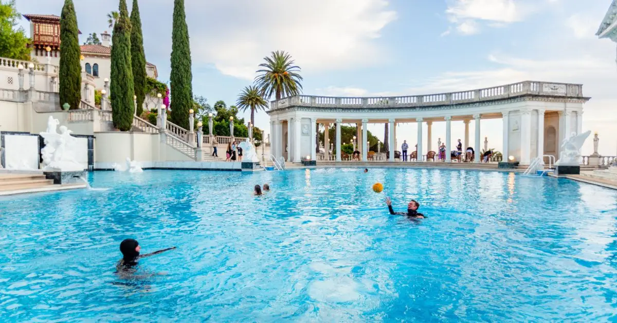 Several people enjoy swimming in one of the best hotel pools in California, surrounded by ornate statues and columns under a clear sky. In the background, tall cypress and palm trees are visible near a grand building with stairs leading up.