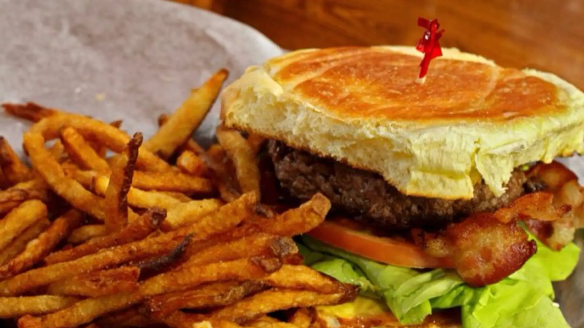 A juicy burger with lettuce, tomato, bacon, and a unique crisscrossed bun is topped with a red toothpick. Known as the best burger on the Big Island, it comes with a generous serving of crispy, golden French fries. The meal is served on a rustic grey plate.