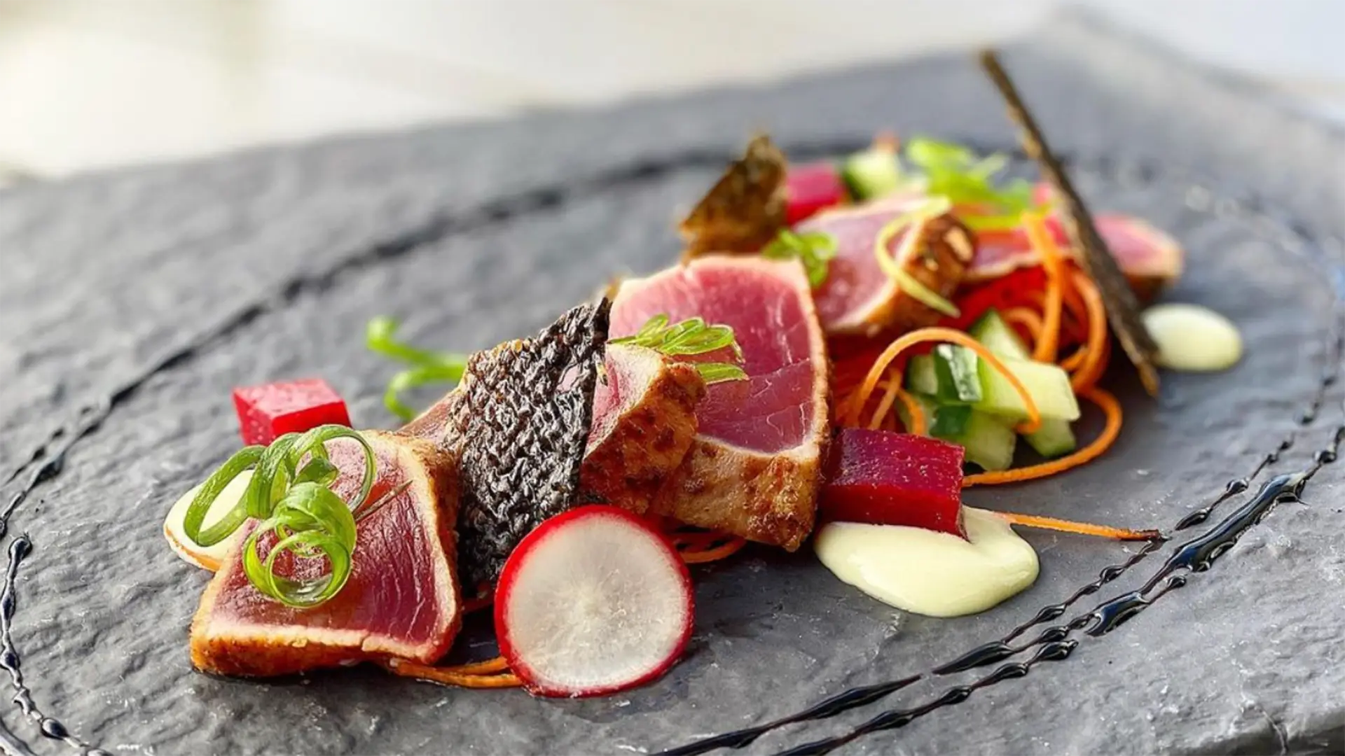 A gourmet dish featuring seared tuna slices, garnished with radish, green onions, and beetroot cubes. The plate is artistically arranged with a drizzle of light green sauce and sits on a textured, dark slate surface reminiscent of Hilo's natural beauty. Small vegetable curls add colorful accents.