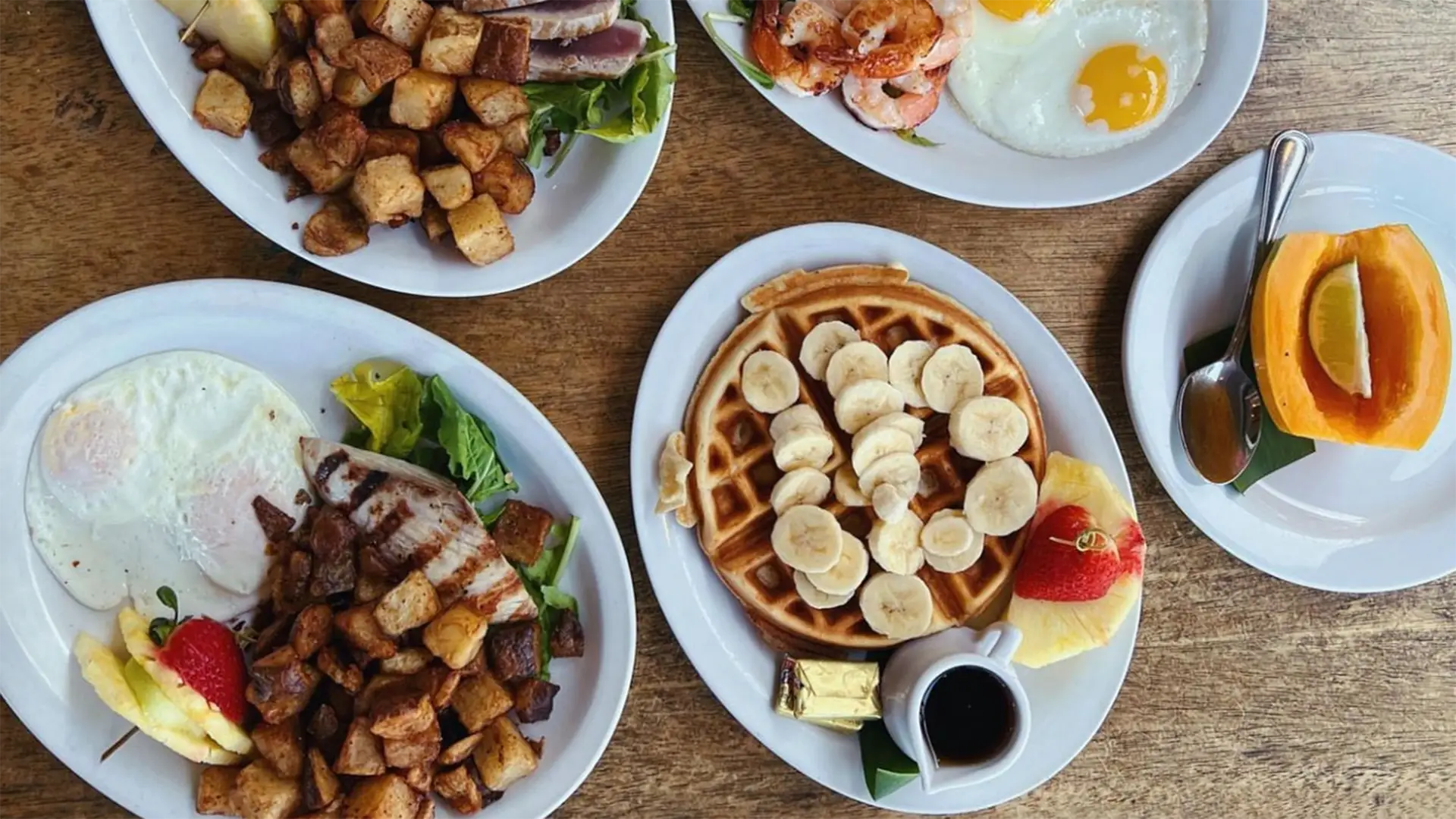 Top-down view of a table with five plates of the best breakfast foods in Kona, including a waffle topped with bananas, home fries, over-easy eggs, toast, a papaya half, and a plate with shrimp. A cup of coffee is also visible.