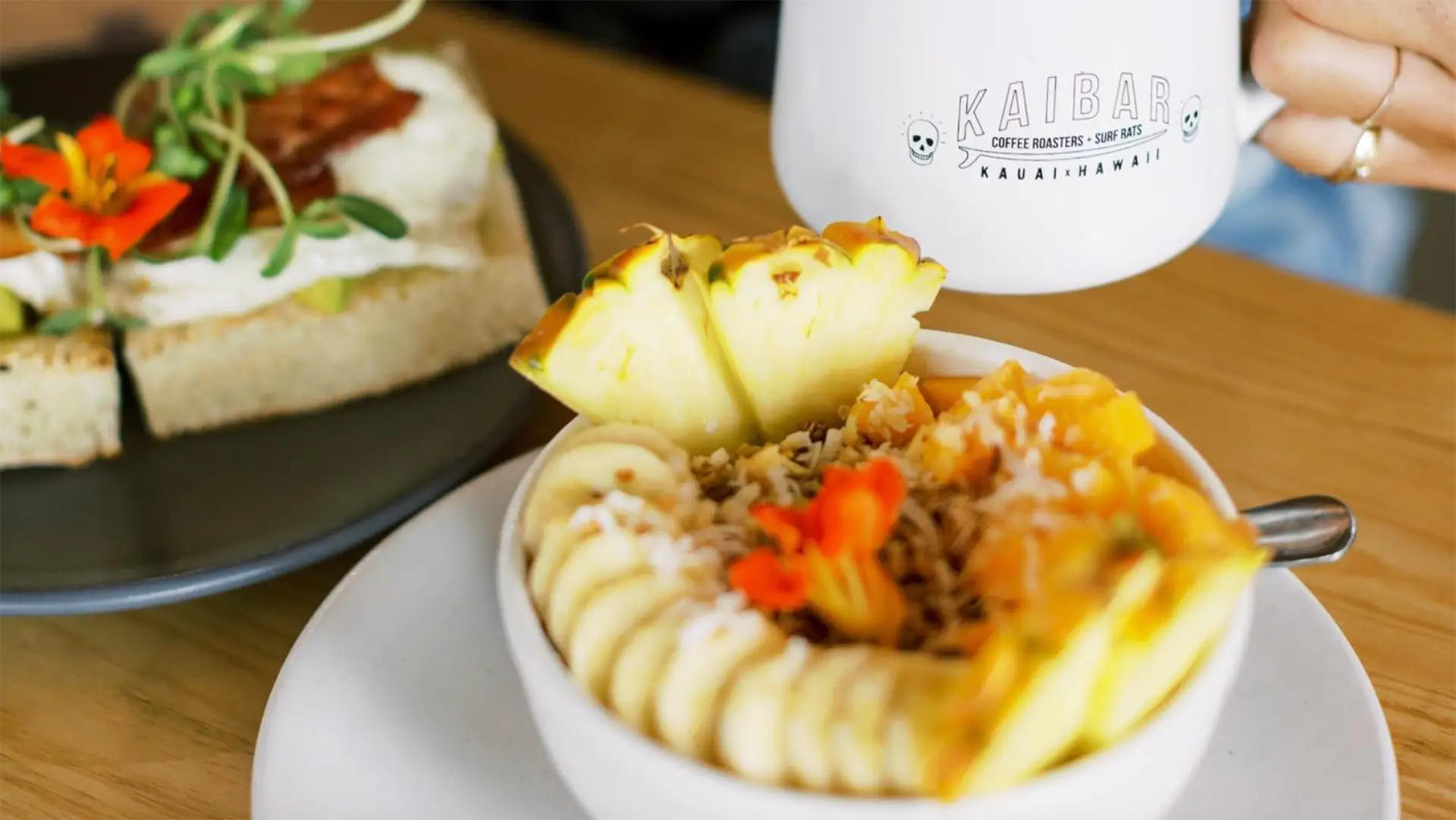 A white bowl filled with sliced bananas, pineapple chunks, and an orange flower is placed on a white plate. A mug labeled "KAIBAK Kauai Hawaii" and a plate with a slice of bread topped with greens and orange flowers are also visible on a wooden table—truly the Best Breakfast in Kauai.