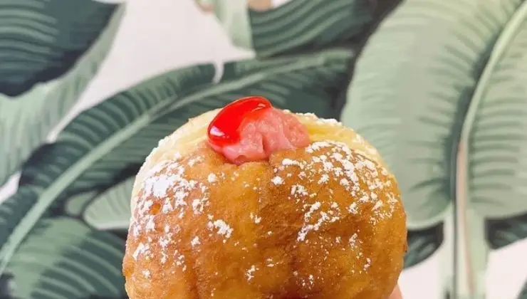 A close-up view of the best malasadas doughnut on Kauai, dusted with powdered sugar and featuring a dollop of red and pink filling on top. The background displays green, tropical leaf-patterned wallpaper.