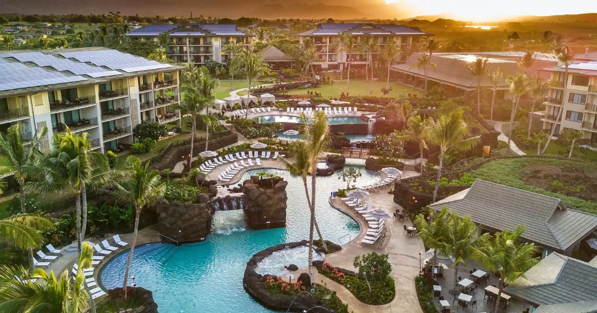Aerial view of a tropical resort at sunset featuring multiple hotel pools surrounded by sun loungers, palm trees, and lush greenery. Multiple buildings with balconies overlook the pool area, and landscaped paths wind through the property.