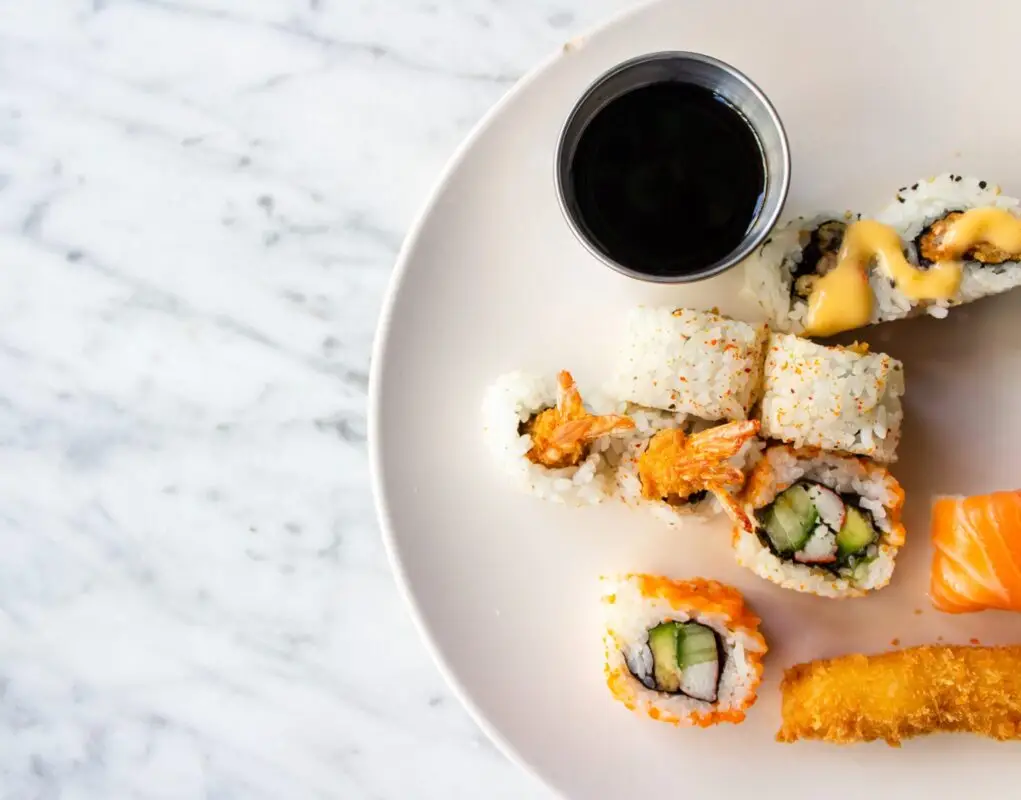 A white plate holding various sushi rolls, including a piece of salmon nigiri, tempura roll, and California roll. There is also a small metal cup filled with soy sauce on the plate. The background is a white marble surface—an image reminiscent of the best sushi near and around Lihue, Kapa'a, Hokuala.