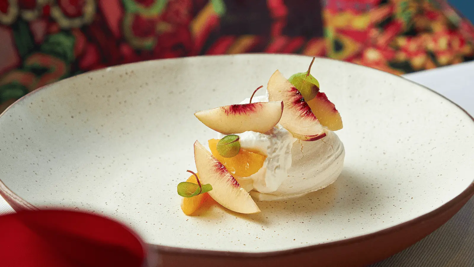 A contender for the Best Desserts in the Bay Area, this gourmet treat features a scoop of vanilla ice cream or sorbet elegantly topped with thin, artfully arranged slices of peach and small green garnish leaves, all served on a minimalist speckled ceramic plate against vibrant, colorful patterns.