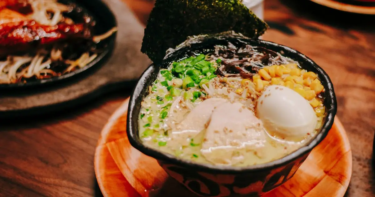 A bowl of ramen, filled with broth, noodles, corn, green onions, seaweed, and slices of chicken or pork, topped with a halved soft-boiled egg. The dish is presented on a wooden surface with another dish visible in the background—truly one of the best lunch spots in San Francisco.