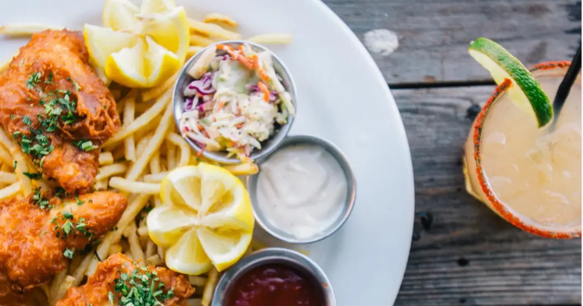 A plate of fish and chips garnished with lemon wedges and a side of coleslaw, tartar sauce, and ketchup. The dish is served on a rustic wooden table next to a beverage with a salted rim and lime garnish—truly the best lunch in San Francisco.