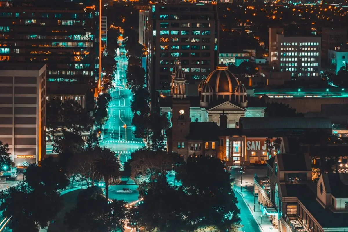Aerial night view of an urban downtown area in the South Bay with illuminated streets and buildings. Notable structures include a clock tower and a domed building in the foreground. The city lights create a vibrant contrast against the dark sky. "SAN JOSE" signage is visible.