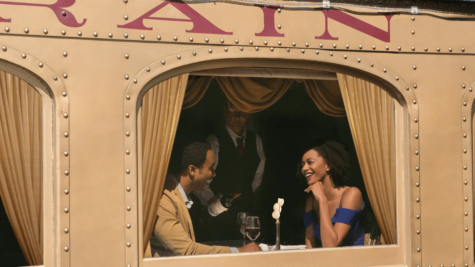 A couple sits at a table inside a vintage-style train carriage, enjoying a meal together. Both are smiling and engaged in conversation. A waiter, dressed in a formal uniform, stands in the background holding a bottle of wine. The setting appears elegant and cozy—one of the best things to do in Napa this April.