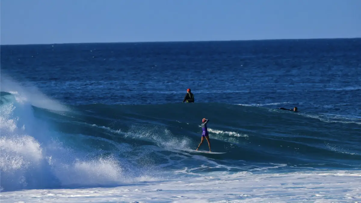 A surfer in a purple wetsuit rides a large wave against a deep blue ocean backdrop, typical of the North Shore annual events. Another surfer in a dark wetsuit floats in the water behind them, with a third person visible further in the background. White foam from breaking waves is in the foreground.